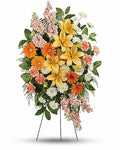 A vibrant floral arrangement featuring yellow lilies, orange daisies, white chrysanthemums, and pink filler flowers with green foliage, displayed on a metal stand against a white background.